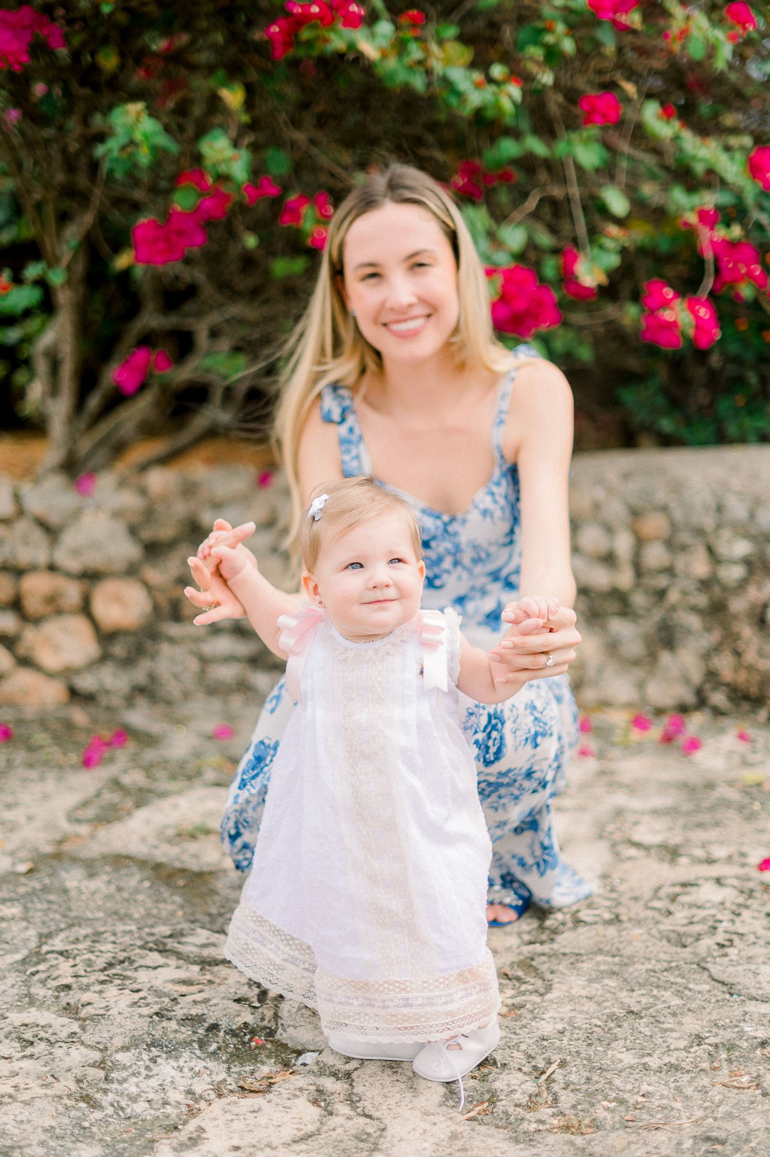 A toddler in a white dress poses with adult by blooming bougainvillea flowers.