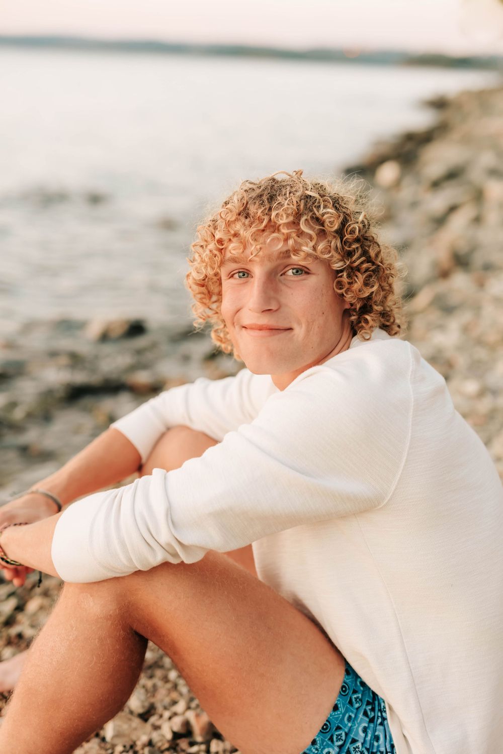 Casual beach portrait of someone in a white sweater sitting on rocky shore at sunset.