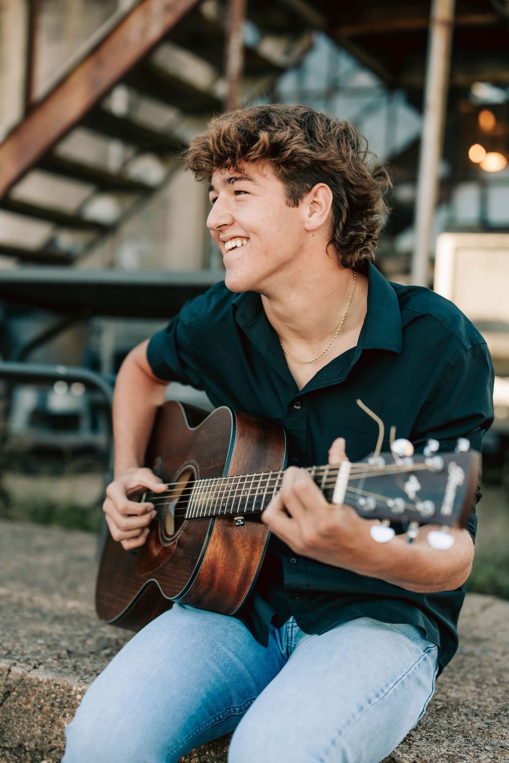 Person plays acoustic guitar while sitting outdoors with industrial backdrop.