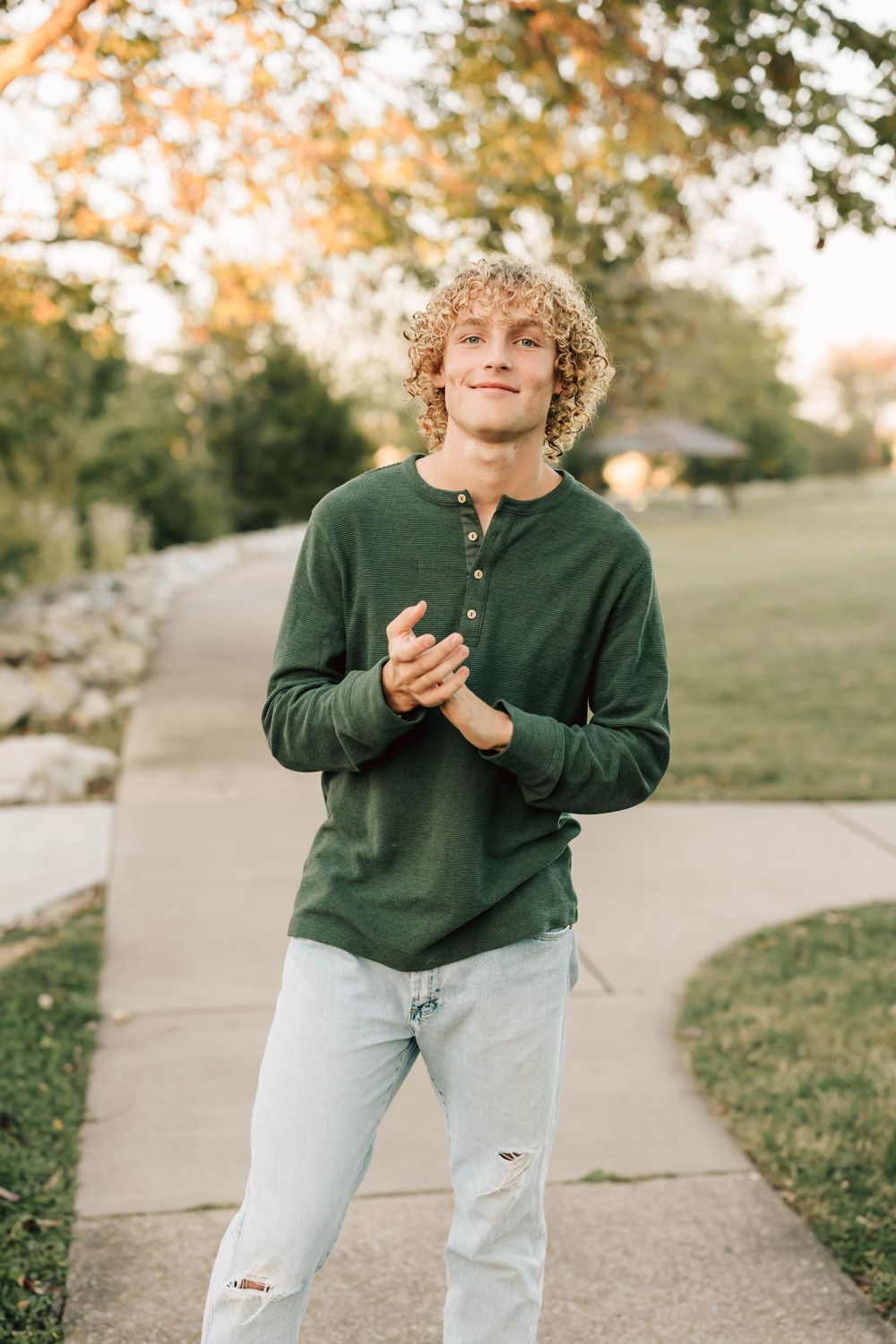Casual portrait of someone in a green henley shirt and ripped jeans standing on a sidewalk during autumn.