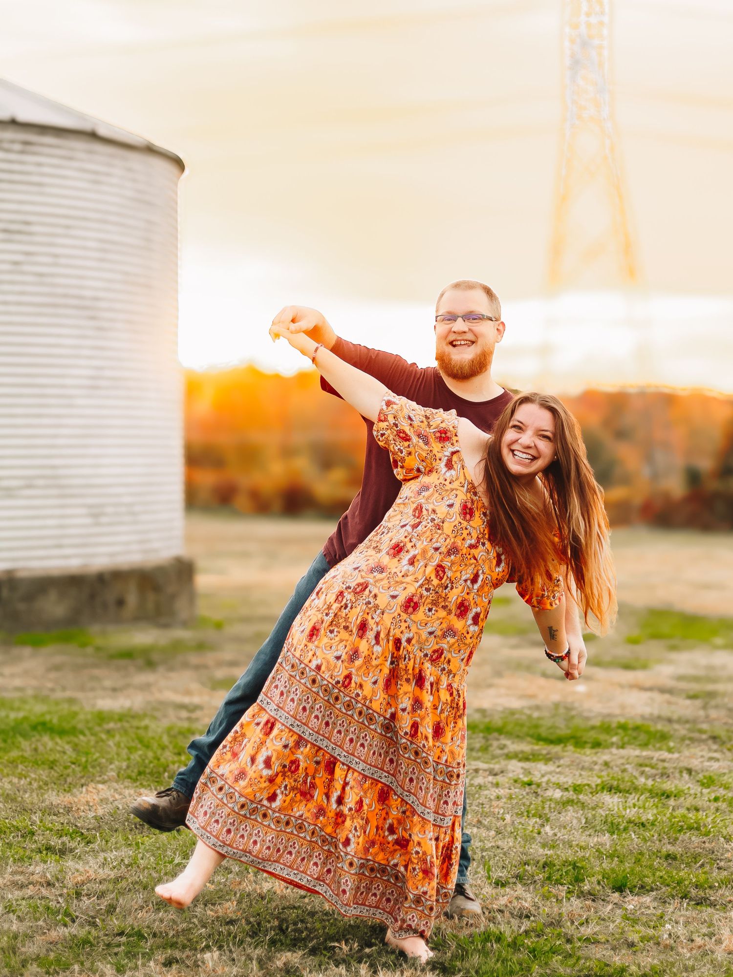 Couple playfully posing in a field at sunset with grain silos and autumn foliage in the background.