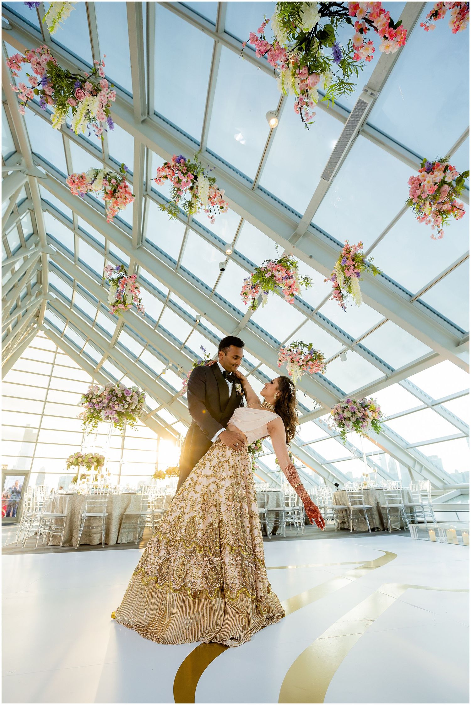 Romantic scene in a glass-ceiling venue with hanging pink floral arrangements and golden floor accents.