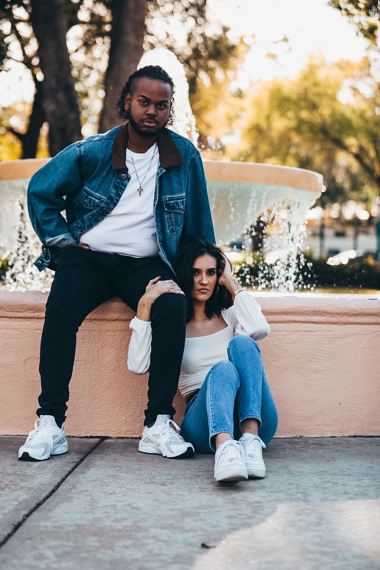 Stylish couple in denim posing near fountain, man standing woman sitting on ground.
