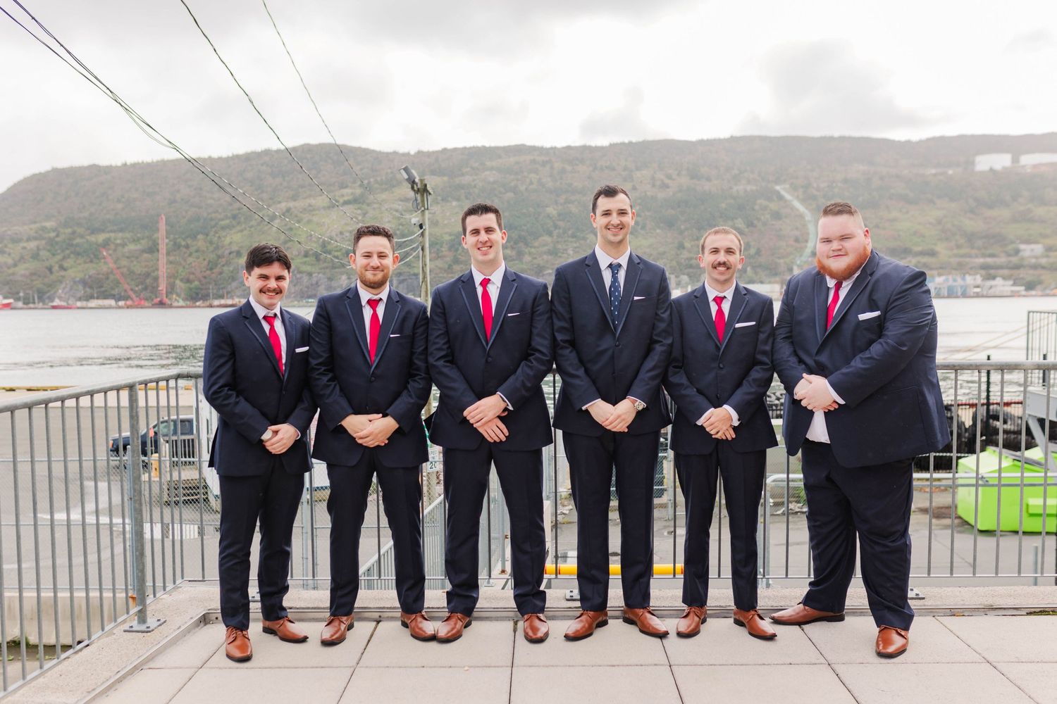 Groomsmen in matching dark suits show off colorful socks while posing on outdoor deck.