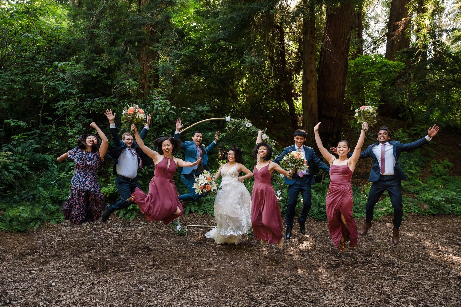 Wedding party jumping joyfully in a forest setting wearing burgundy and navy attire.