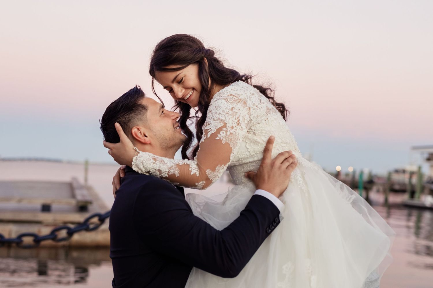 Romantic wedding couple embraces at sunset with bride in white lace dress and groom in dark suit.