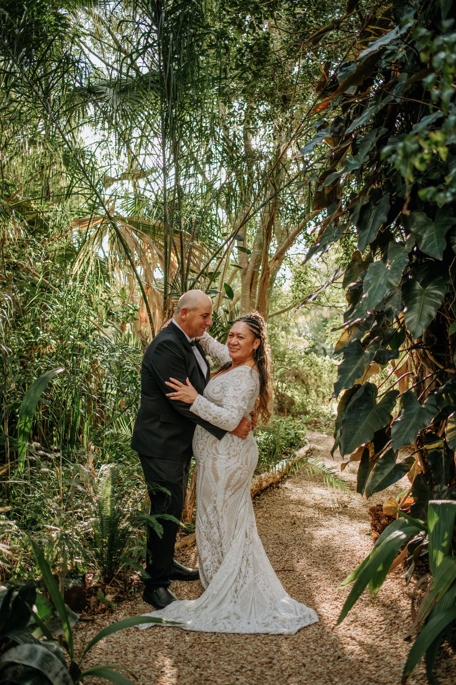 Wedding couple poses on woodland path in elegant attire surrounded by tropical garden scenery.