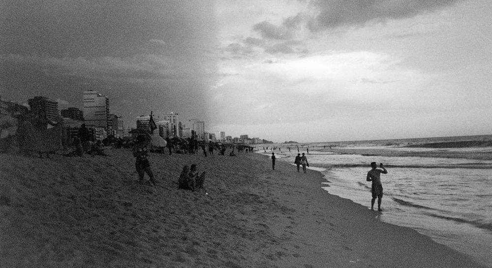 Black and white photograph of beachgoers along a shoreline with city buildings in the background.