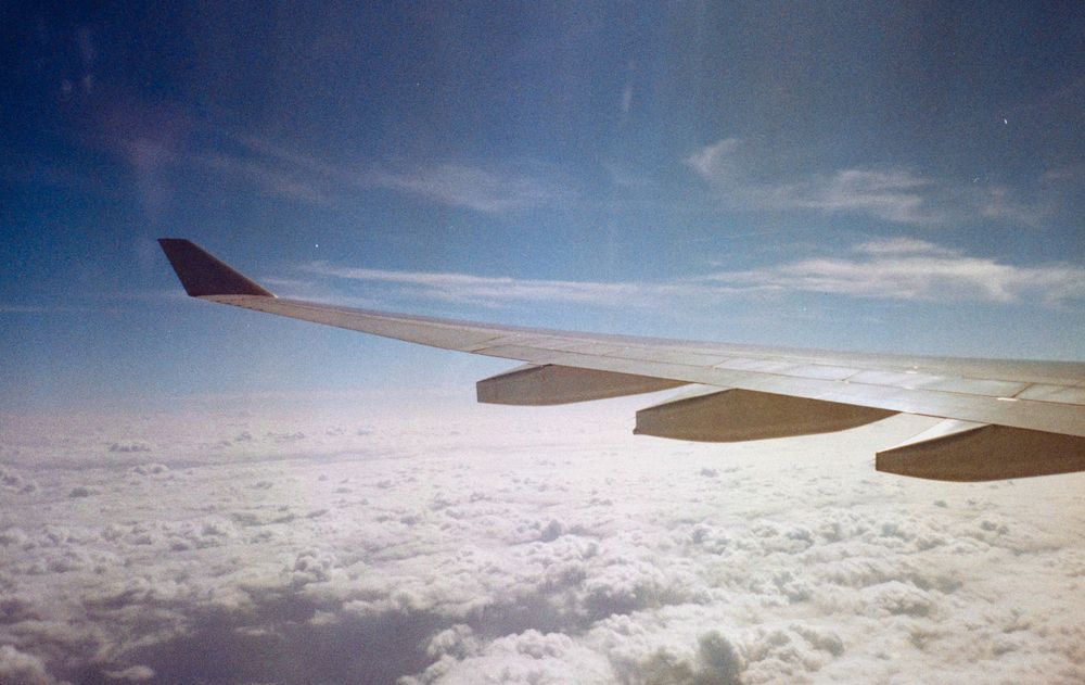 Airplane wing stretches over a sea of white clouds against a bright blue sky.