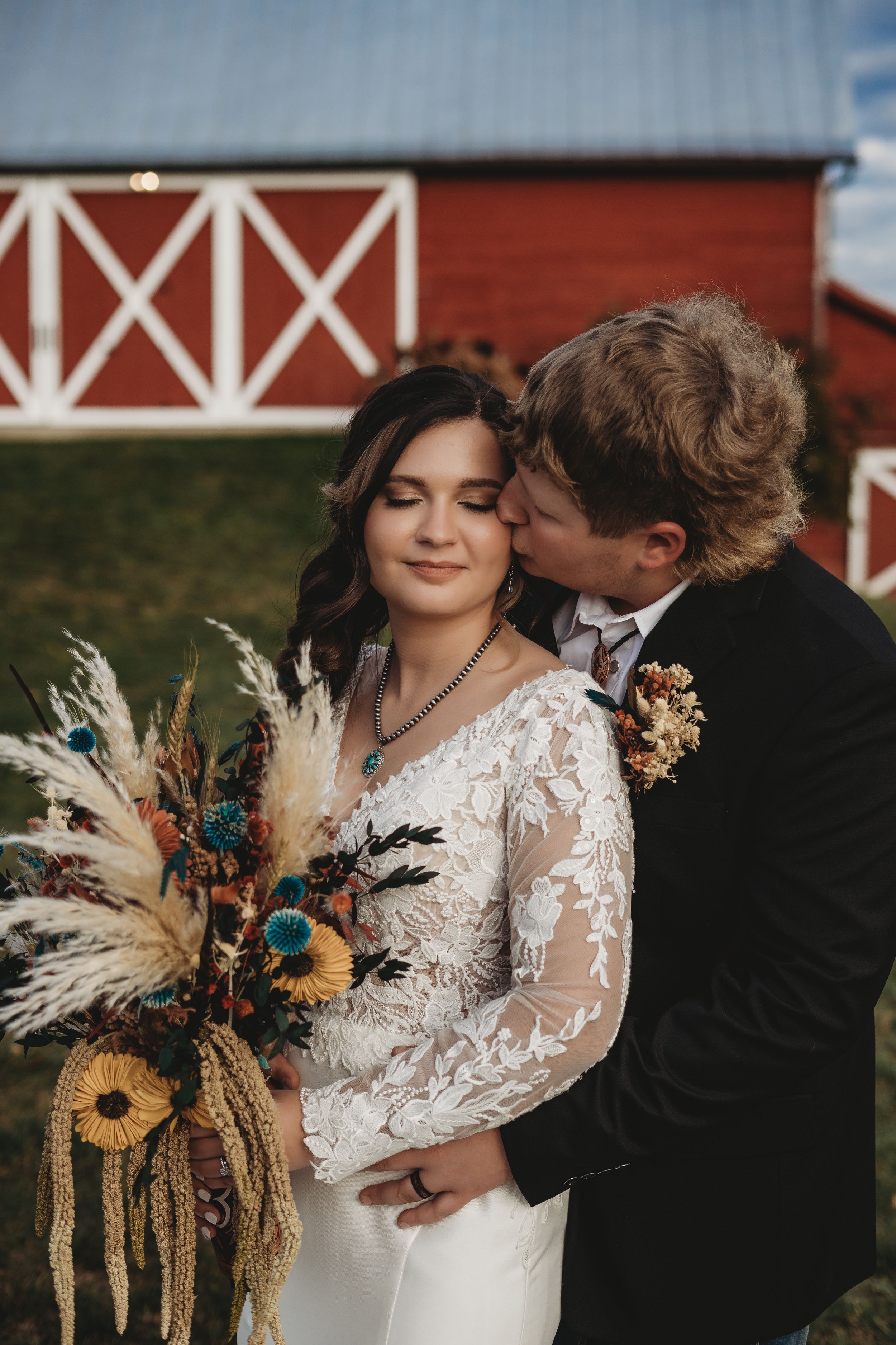 Wedding portraits by red barn featuring dried flower bouquet with sunflowers and wheat stems.