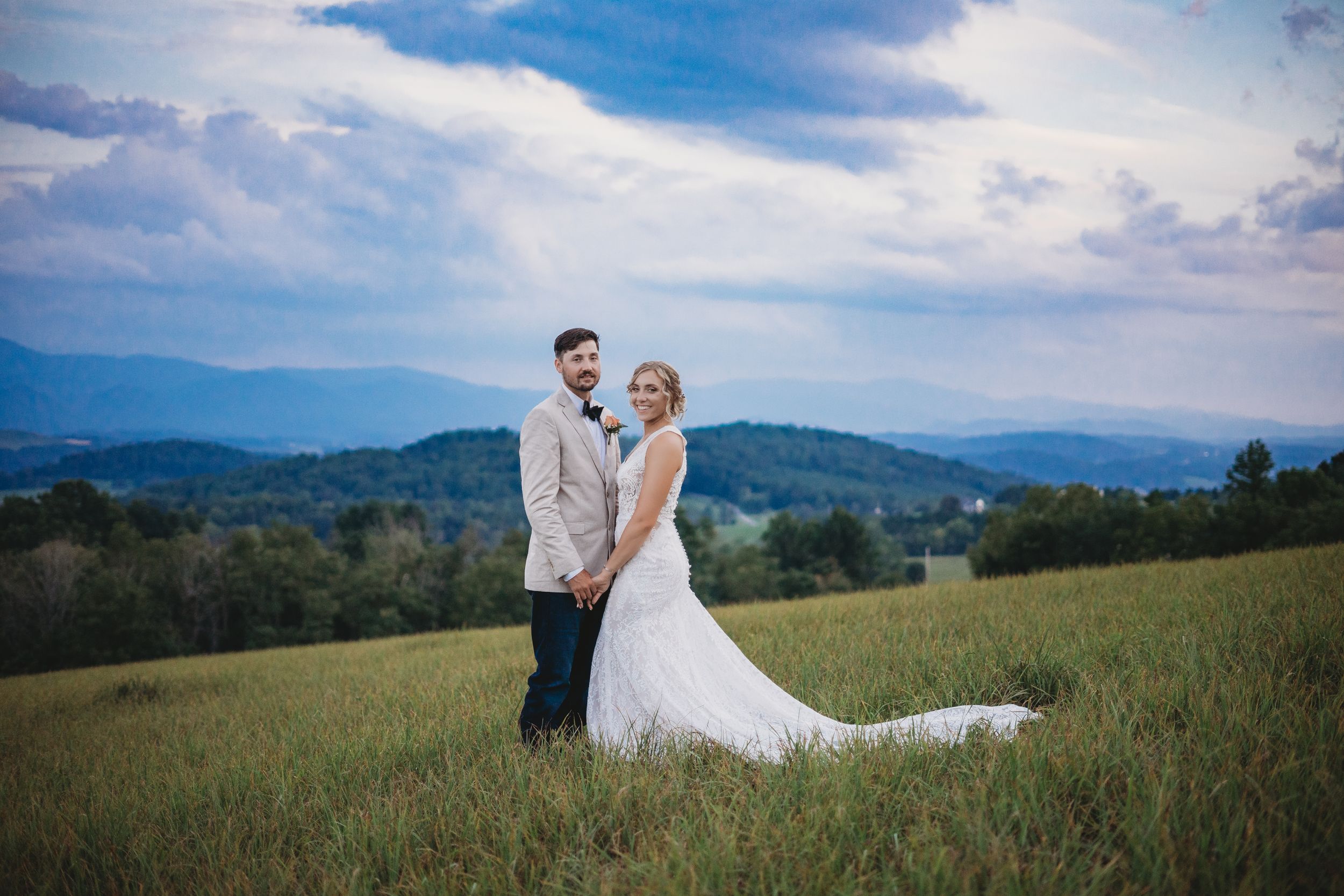 Wedding couple poses in scenic mountain meadow with dramatic blue sky and rolling hills.