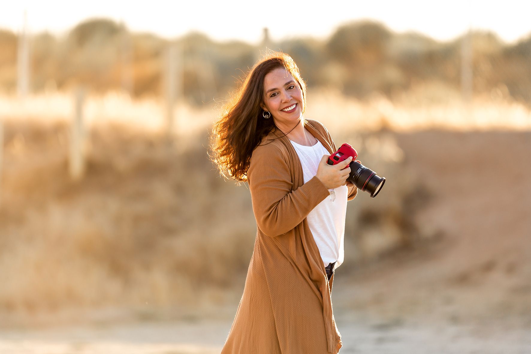A person in a tan cardigan holding a camera outdoors during golden hour.