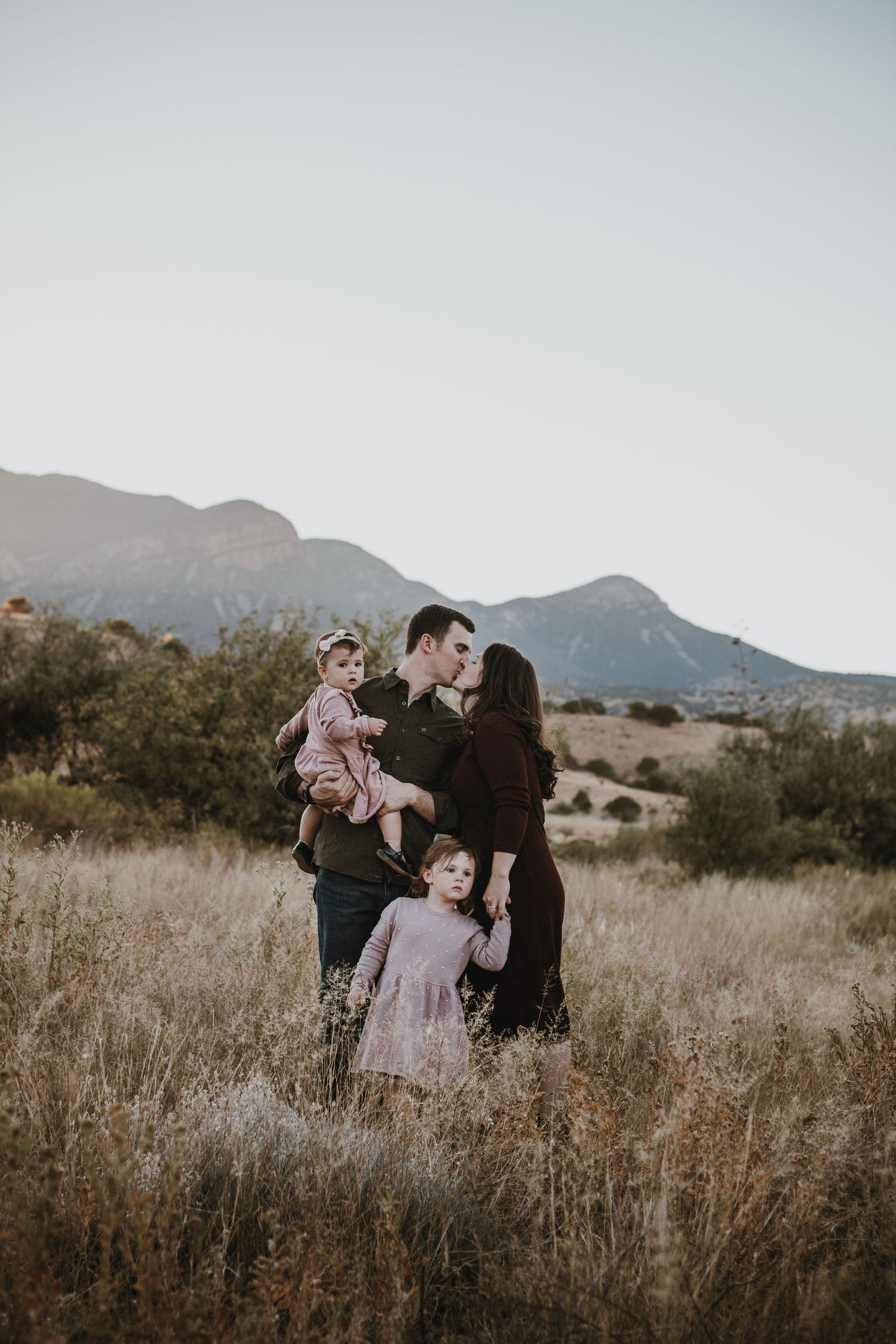mom and dad kissing while holding toddler and holding hands with daughter with huachuca mountains Family photographers kansas city