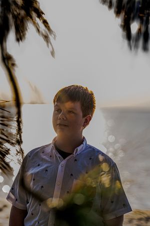 Portrait shot of youth in patterned shirt with palm trees and ocean in background.