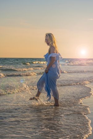A person in a flowing light blue dress walks along the beach shoreline during a golden sunset.