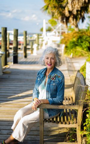 Person in denim jacket sits smiling on wooden bench along coastal boardwalk.