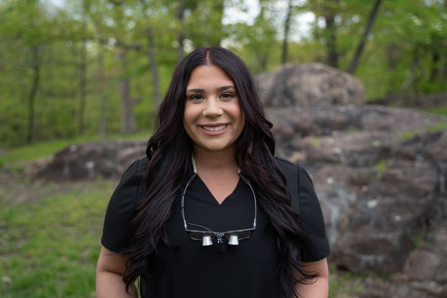 Graduate in black attire posing outdoors against natural rock formations and green trees.