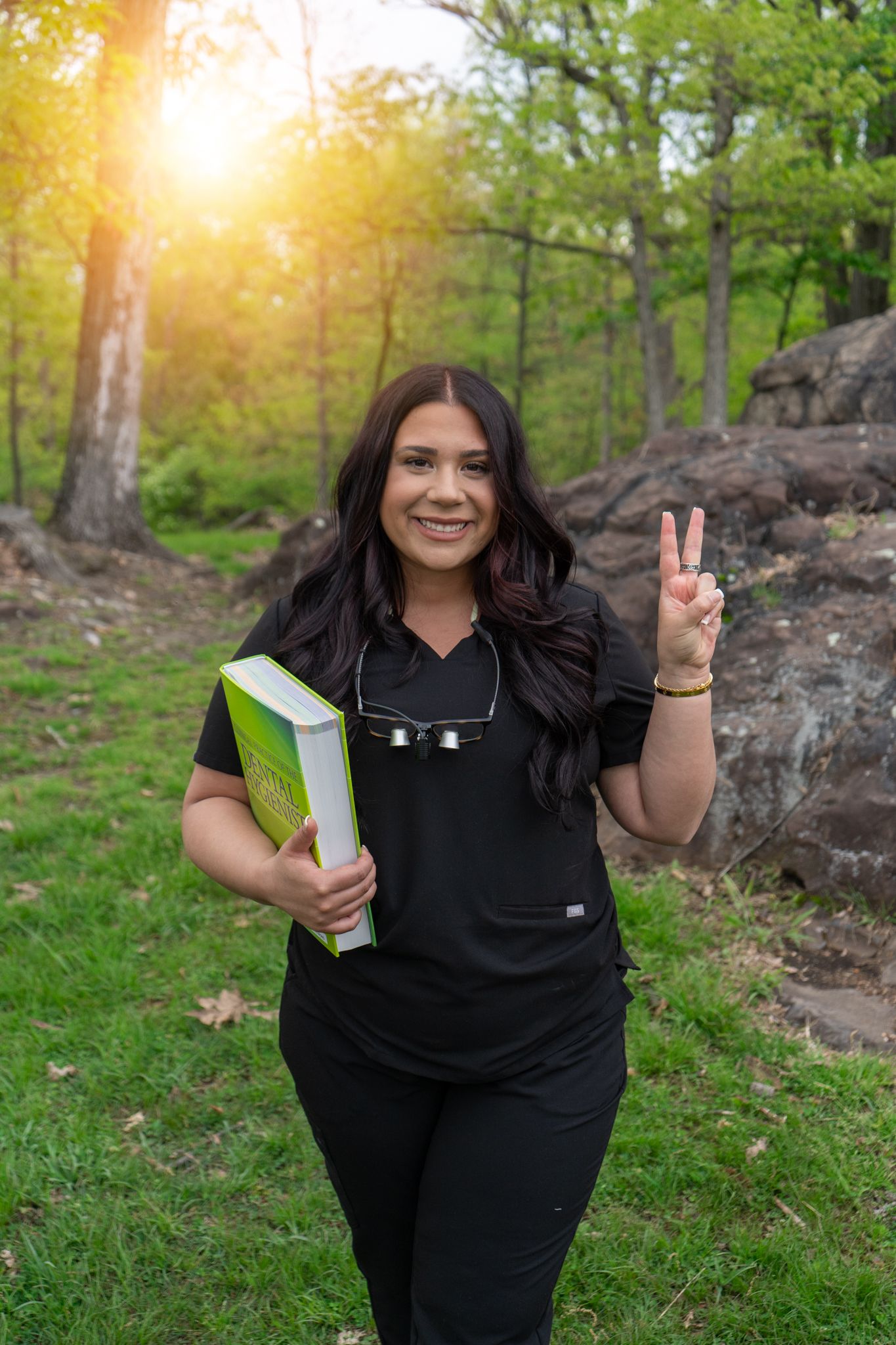 Graduate in black cap and gown making peace sign in sunlit forest setting.
