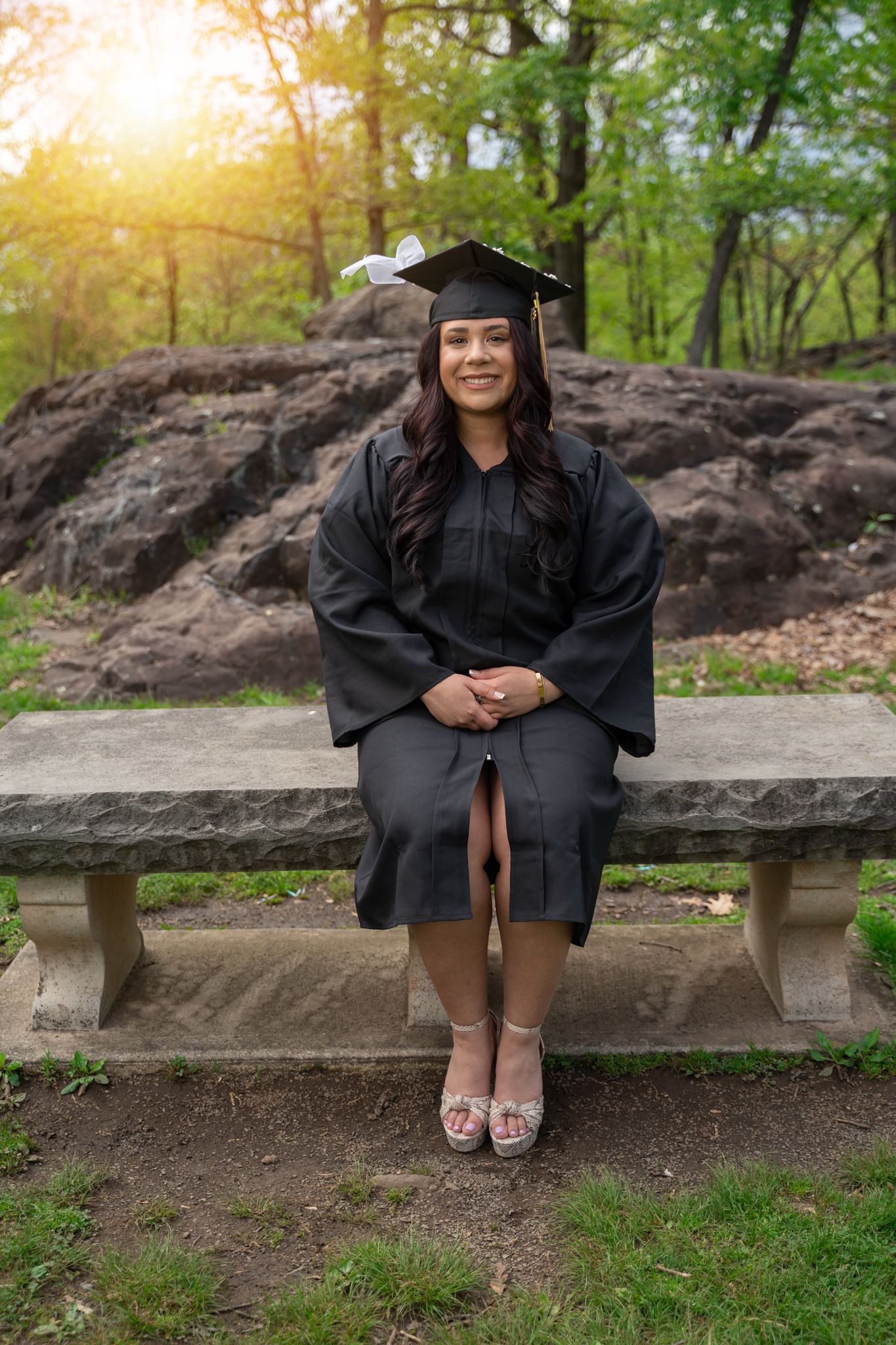 Graduate in black cap and gown sitting on stone bench in wooded park setting.