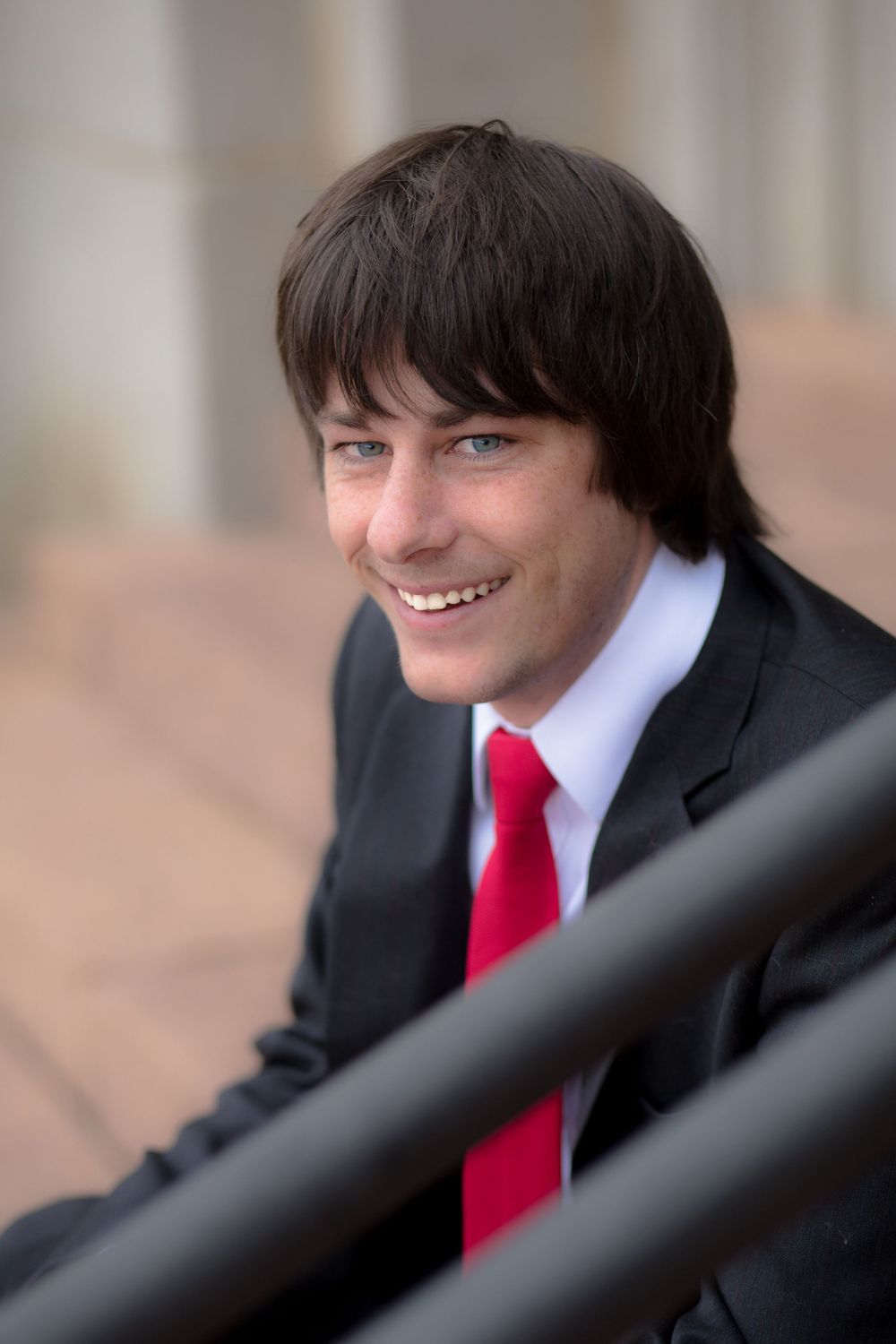 Young man in black suit and red tie smiles warmly at camera.