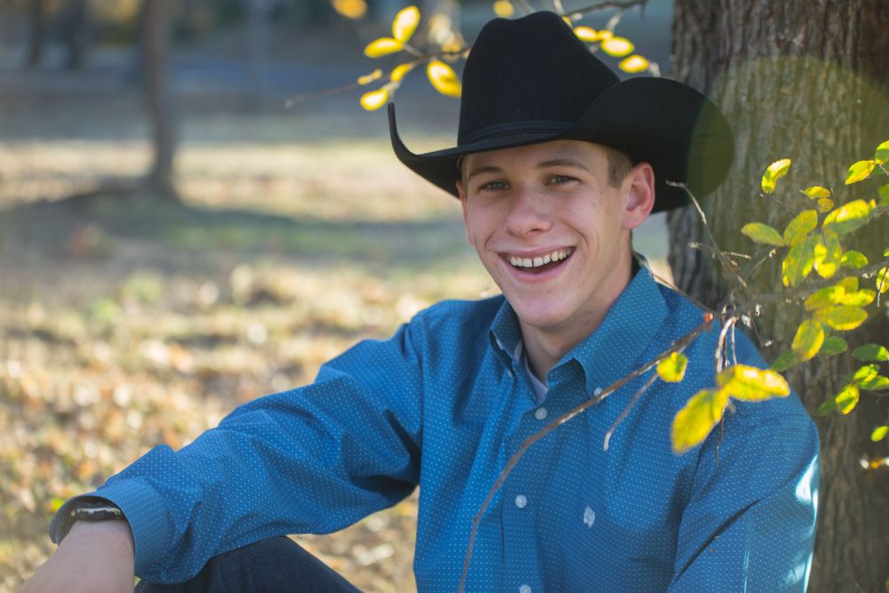 Casual outdoor portrait in cowboy hat and blue shirt sitting by autumn tree.
