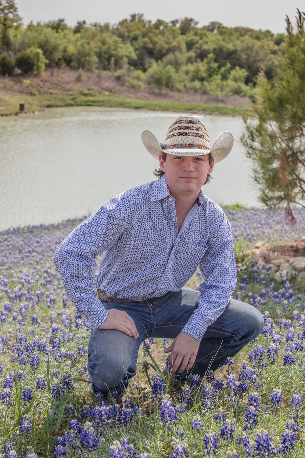 Person wearing western clothes kneeling among bluebonnet wildflowers by a lake.