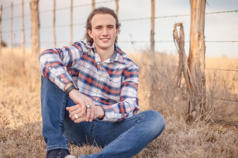 Person in plaid shirt and jeans sits casually in outdoor setting with wooden fence and dry grass.