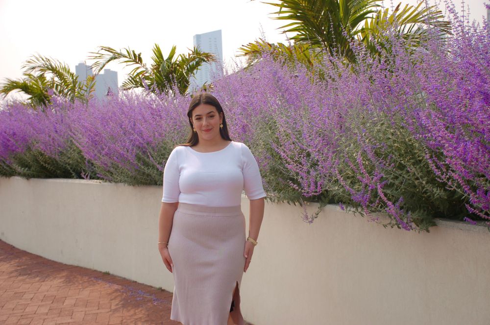 Woman in white dress standing in front of purple flowers and palm trees.