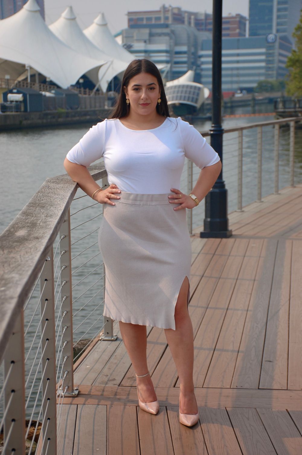 Woman in white top and beige skirt posing on wooden dock with city skyline in background.