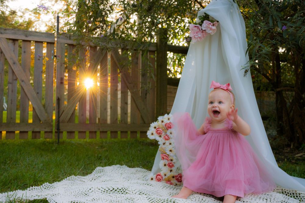 Baby in pink dress sitting under floral canopy with sunlight peeking through fence.