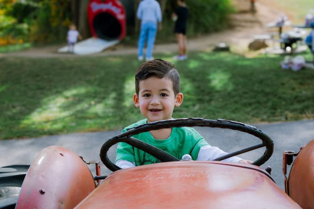 Young boy smiling while sitting in the driver's seat of a pink toy car.