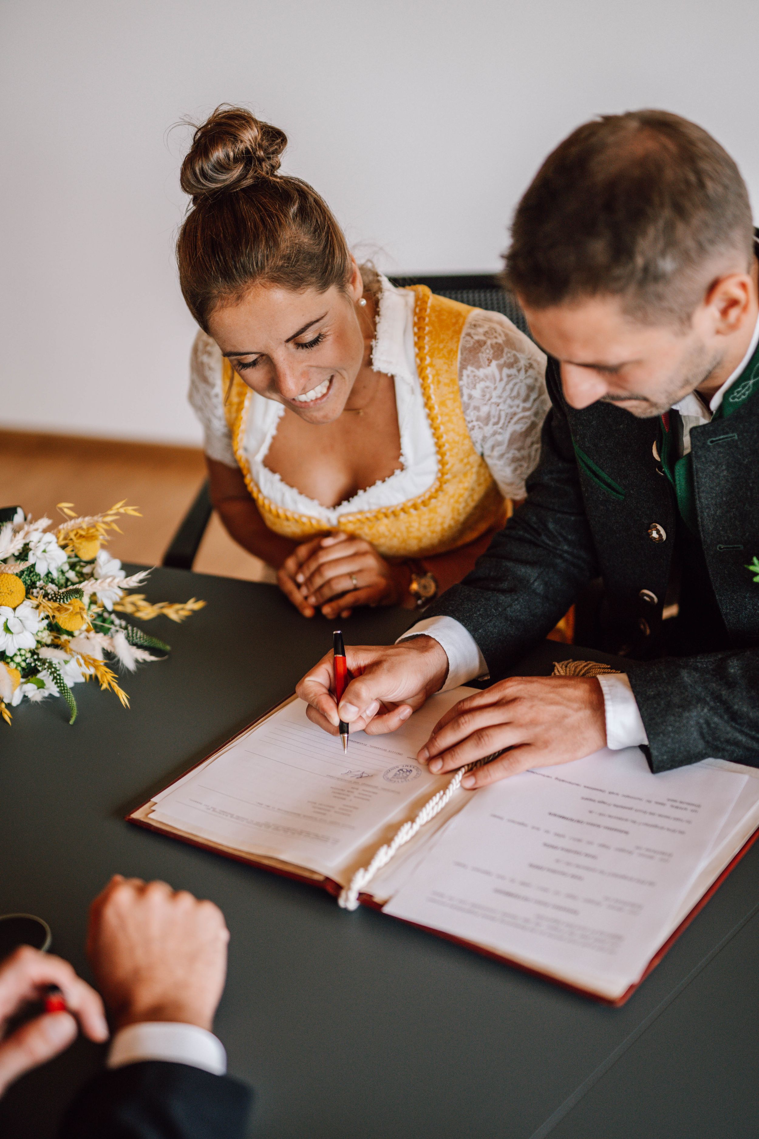 Couple signing marriage documents together at a wooden table with floral decorations.
