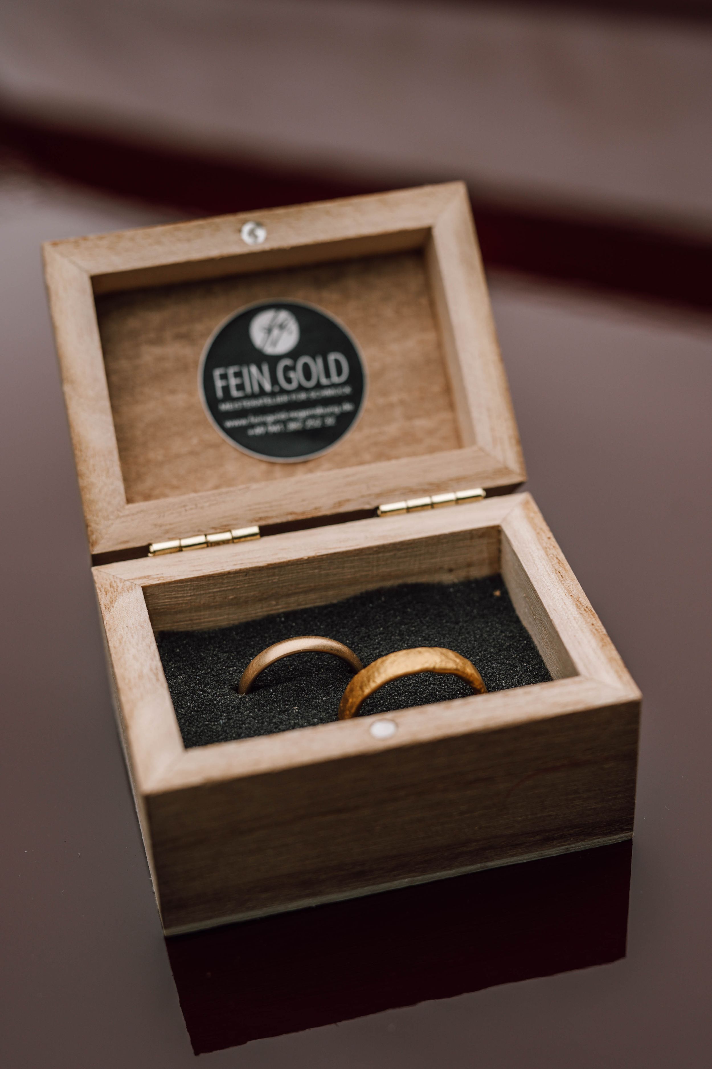 Wooden ring box with 'Fein.Gold' branding containing wedding bands on dark background.