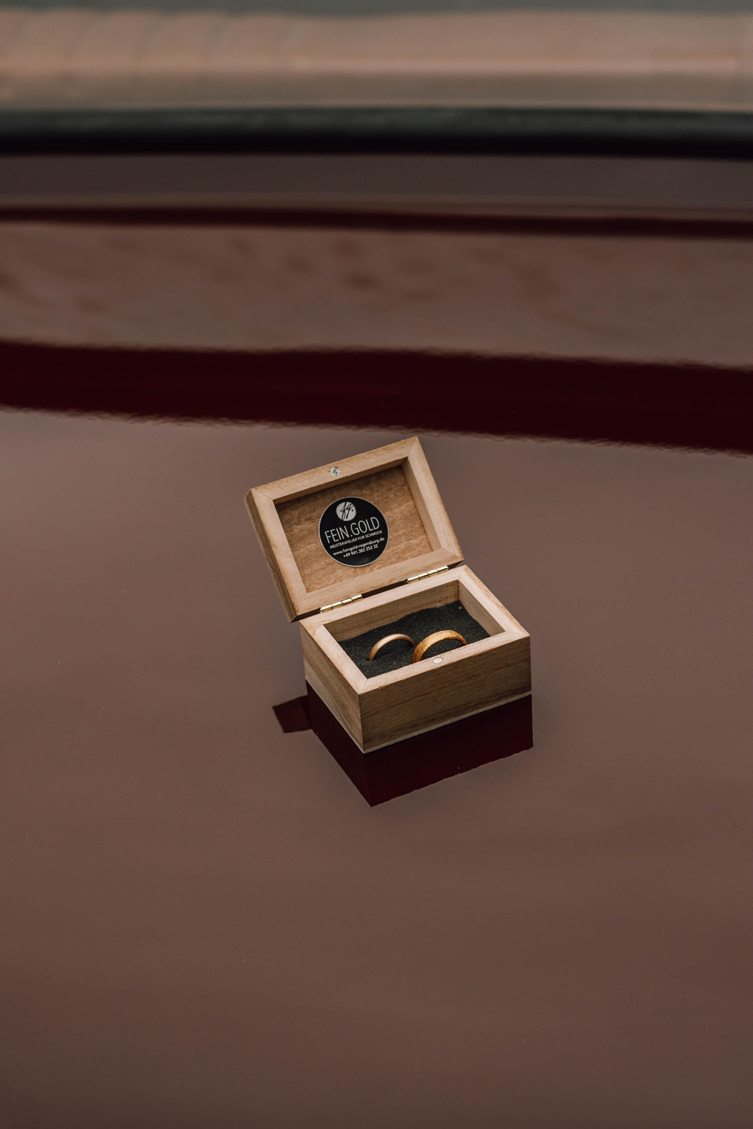 A wooden ring box containing wedding bands sits on a dark surface.