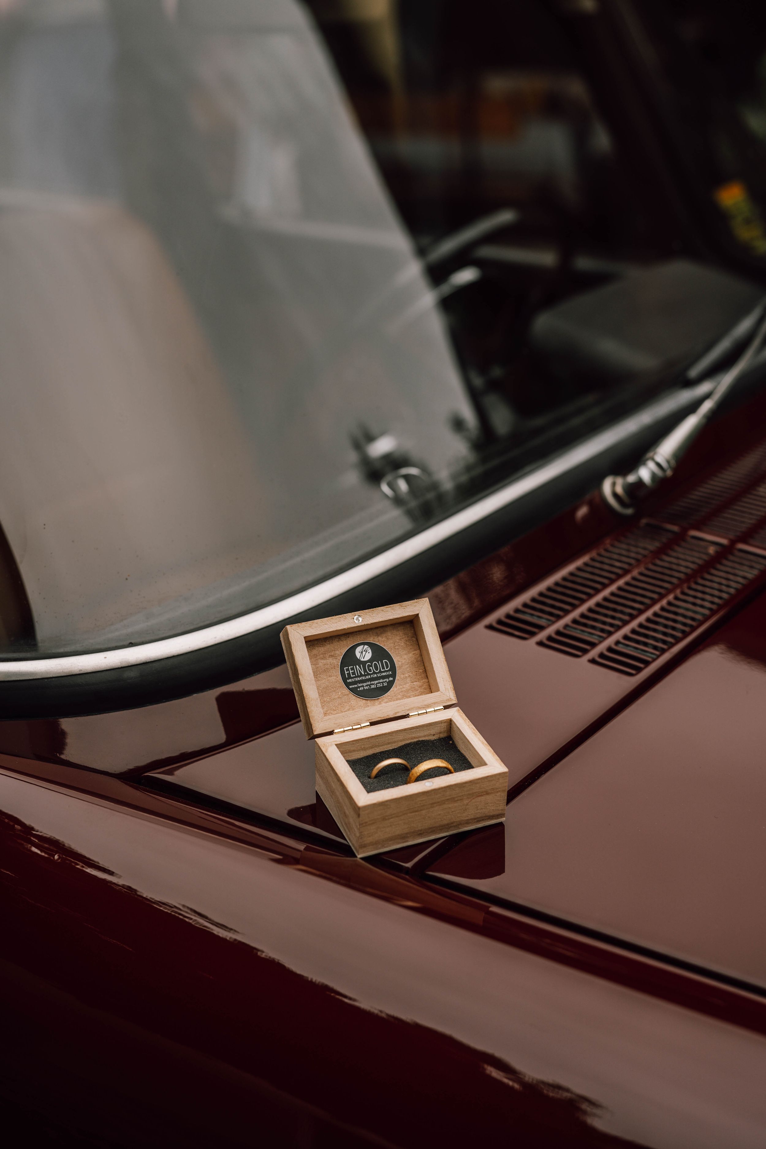 A wooden ring box sits on the hood of a burgundy colored car.