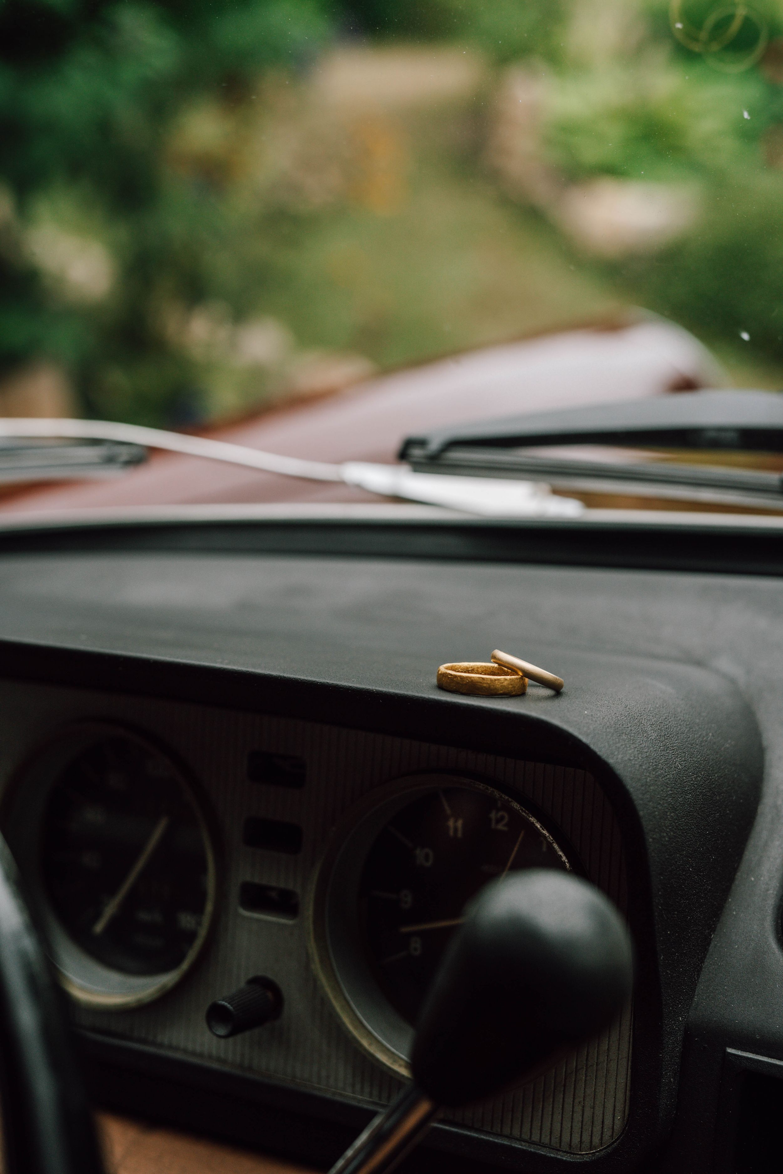 Gold wedding rings placed on black car dashboard with blurred green background.