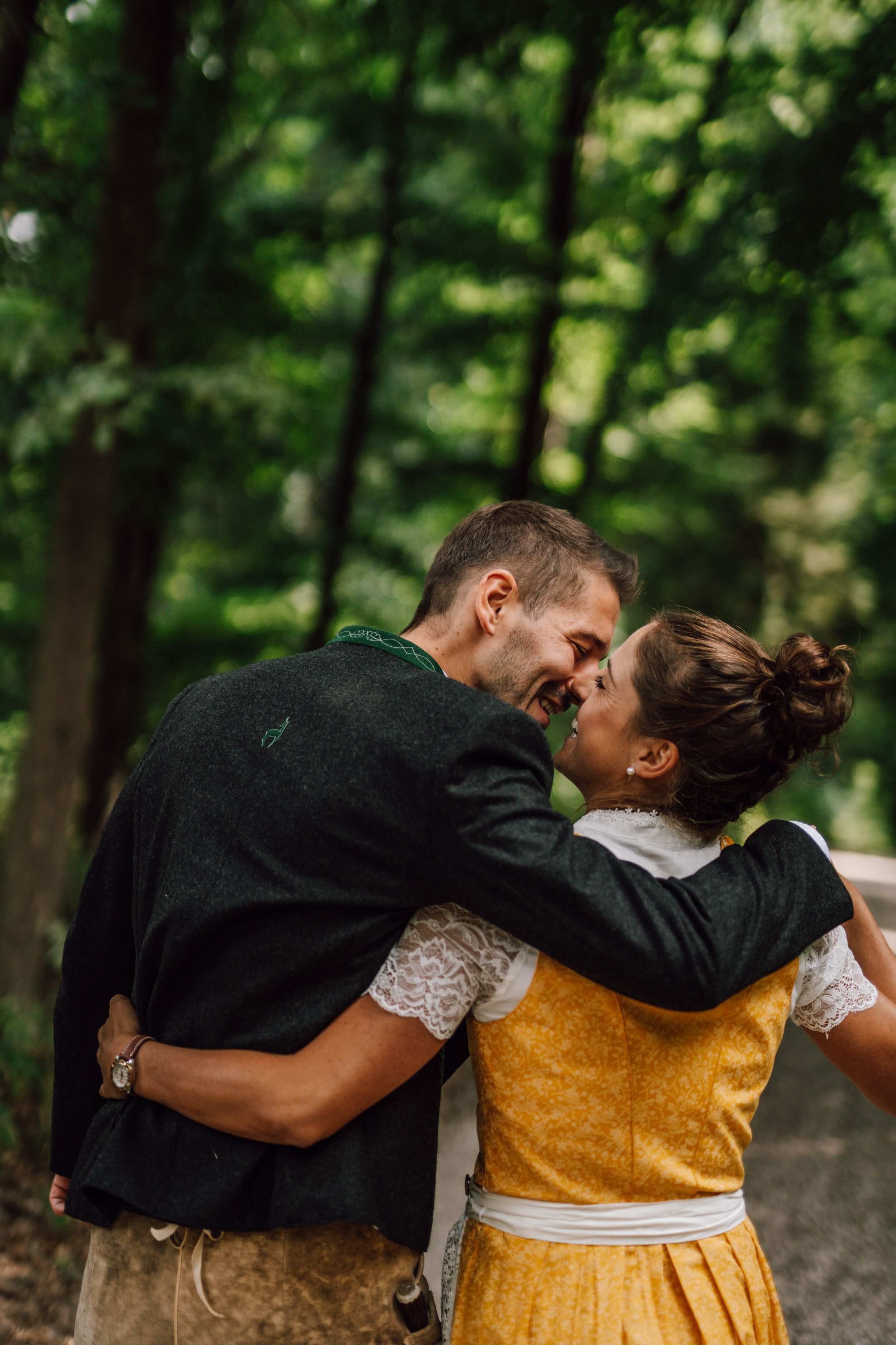 A couple in yellow dirndl and black suit embracing on a wooded path.