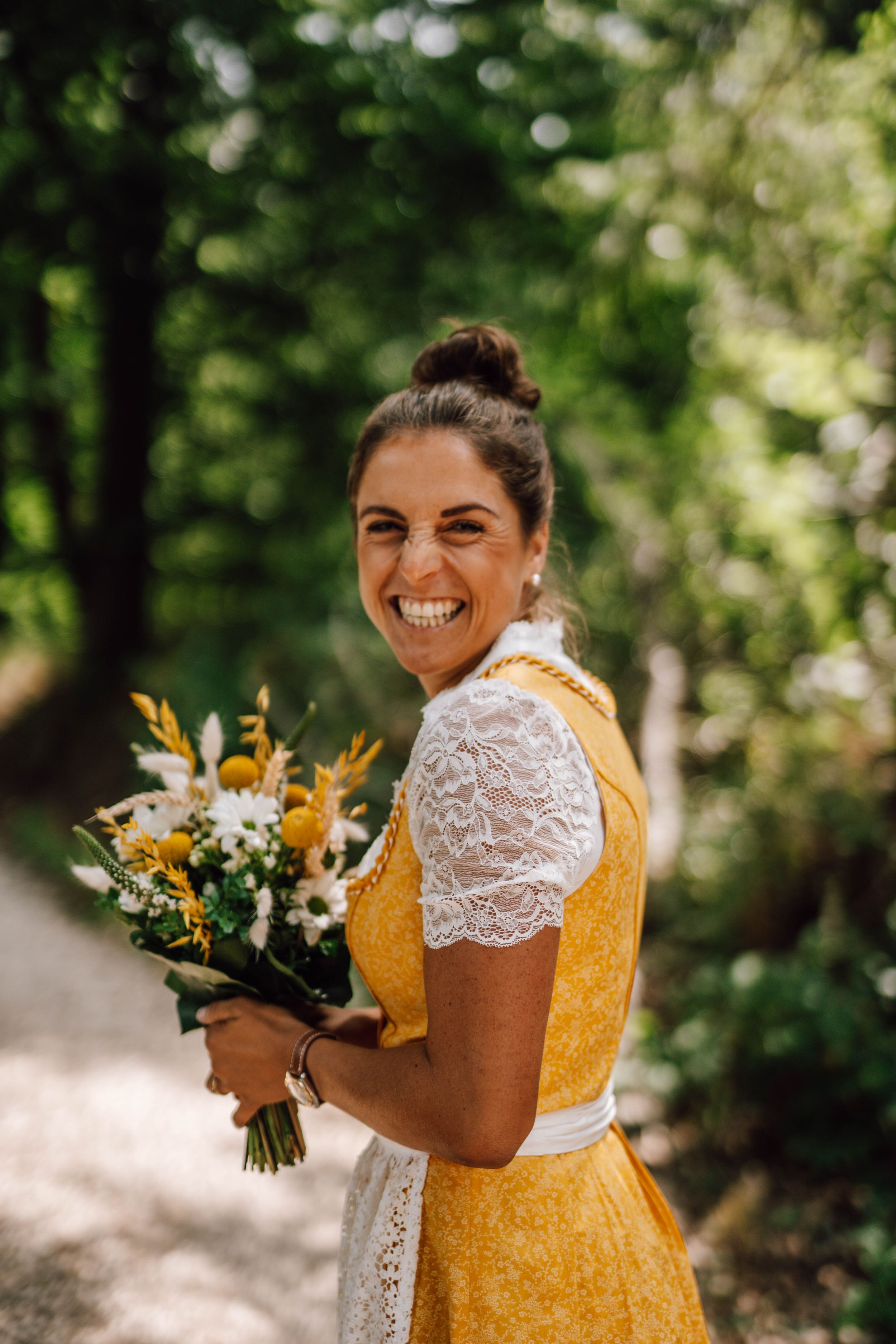 A bride in a yellow dress holds a bouquet of sunflowers and white flowers while smiling.