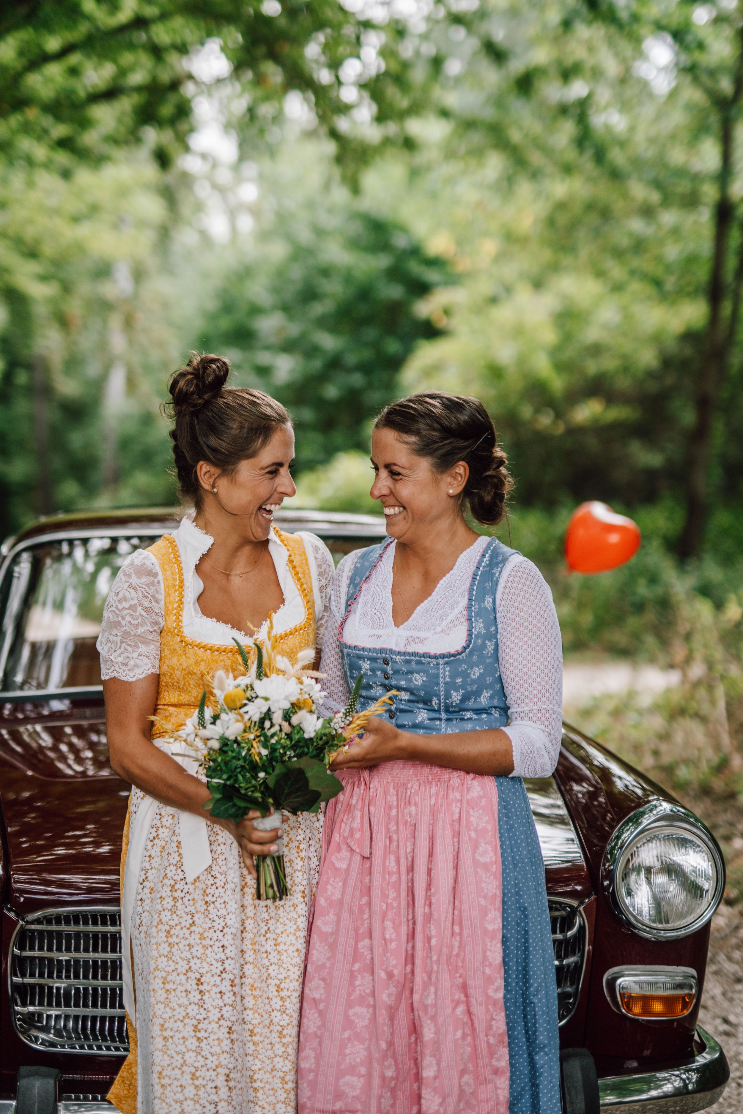 Traditional Bavarian dirndl dresses shown near vintage car in garden setting.