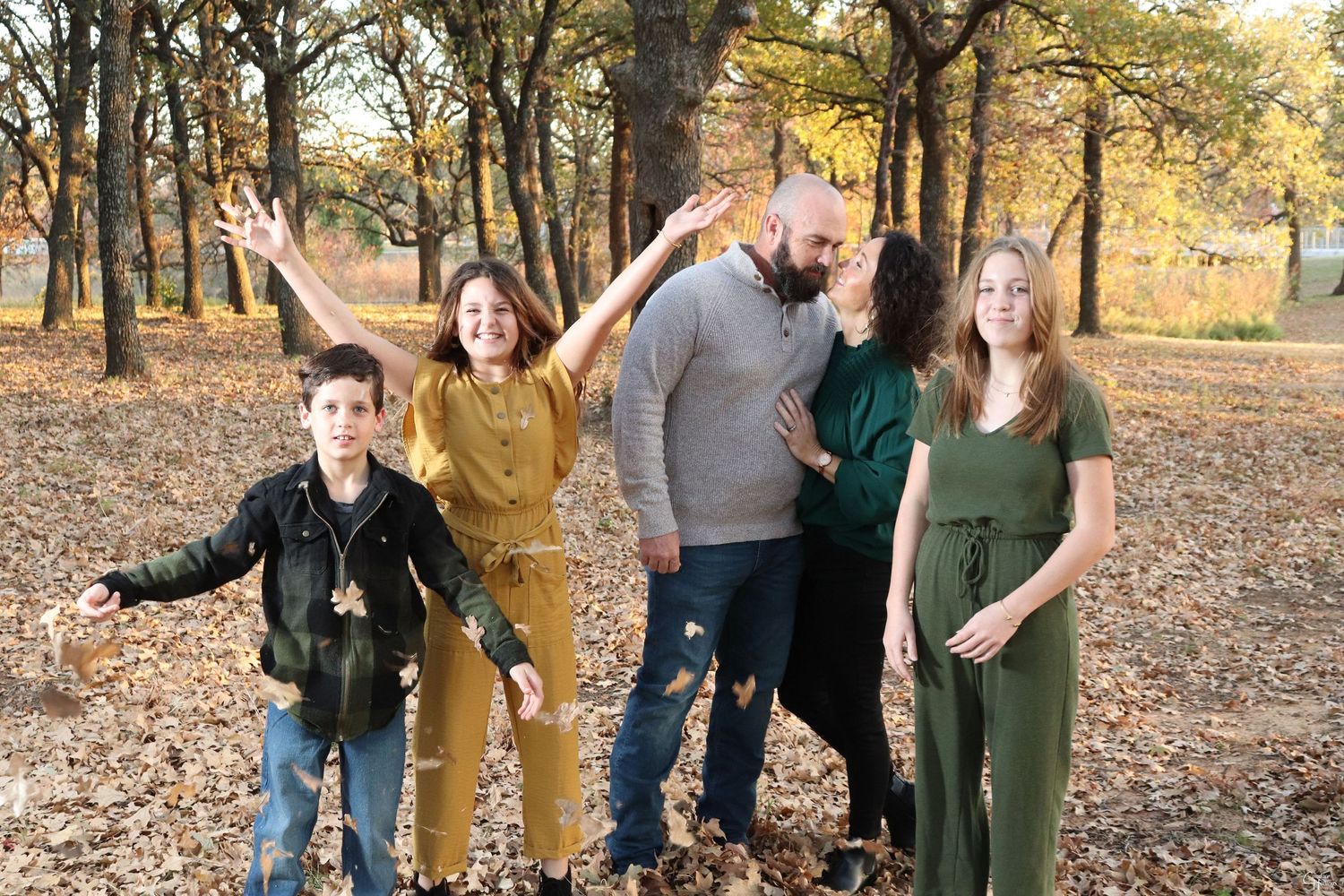 Family in coordinating earth-tone outfits poses playfully among fall leaves in a wooded setting.