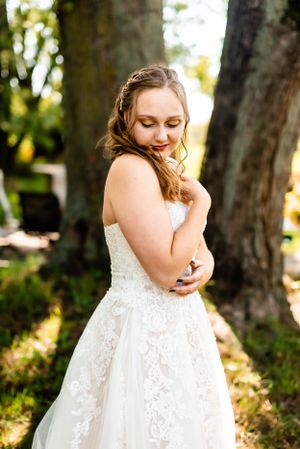 A bride in a stunning lace wedding gown poses gracefully in dappled sunlight among trees.