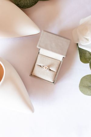 Delicate engagement ring displayed in a beige box with eucalyptus leaves and white flowers.