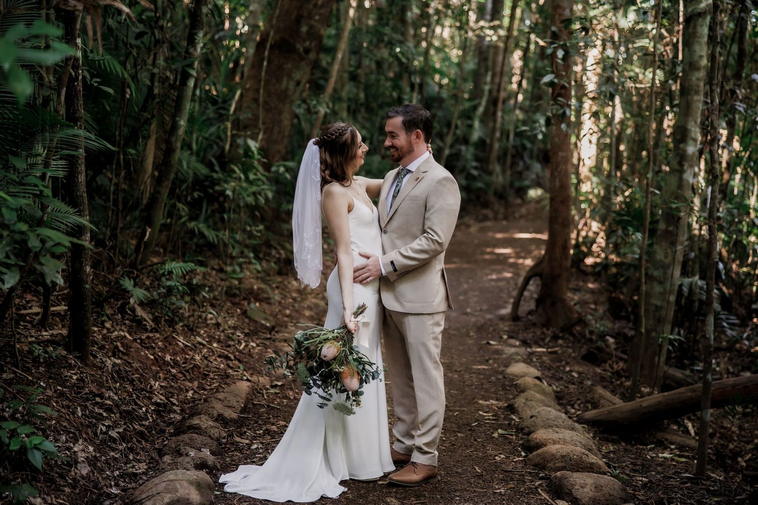 Wedding couple stands intimately on forest path in elegant attire.