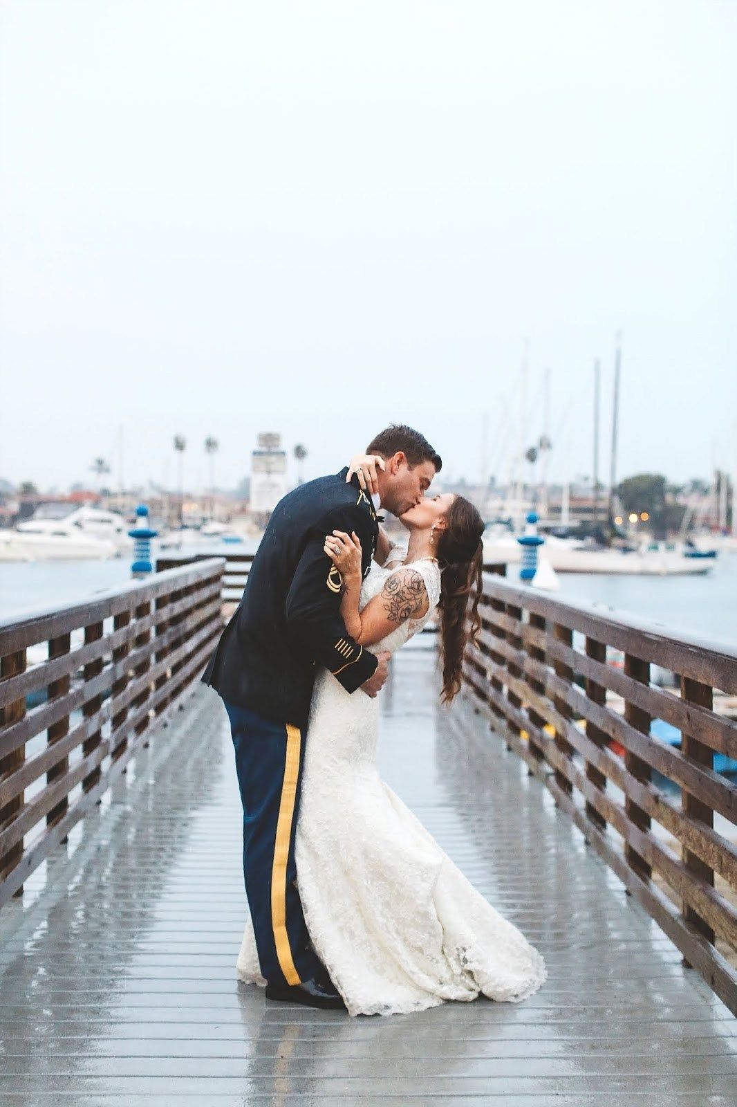 A romantic wedding couple shares a kiss on a wooden pier with boats and palm trees in the background.