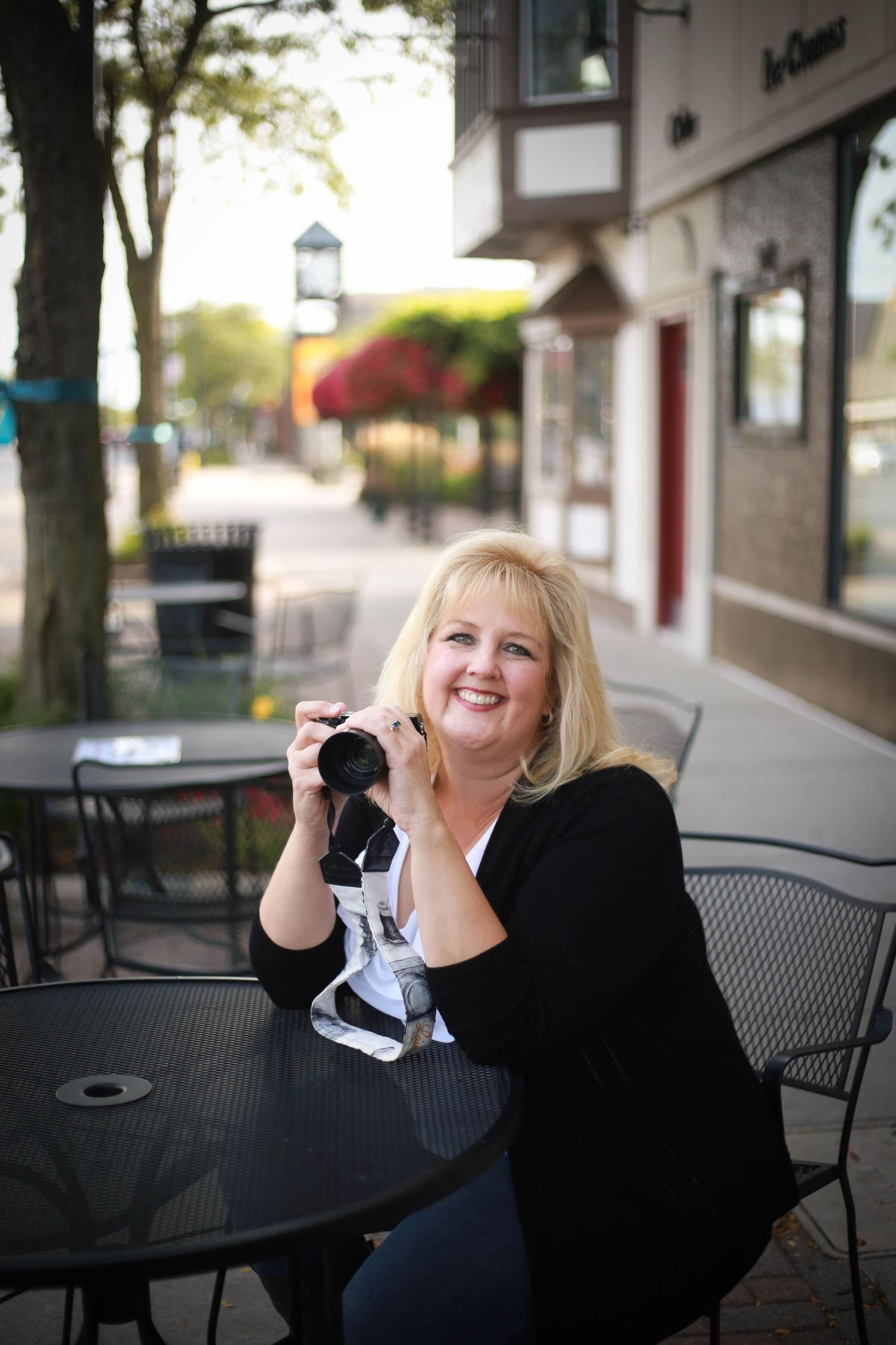 Professional photographer with blonde hair sits at outdoor cafe table holding camera in dark outfit.