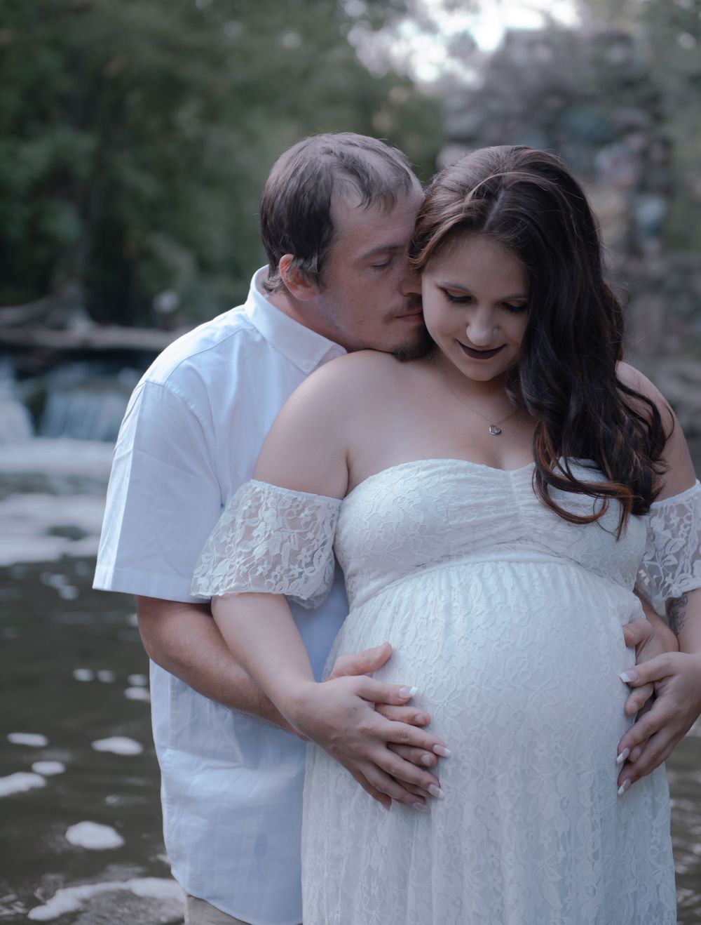 A loving maternity photo showing a couple in white embracing by a stream.