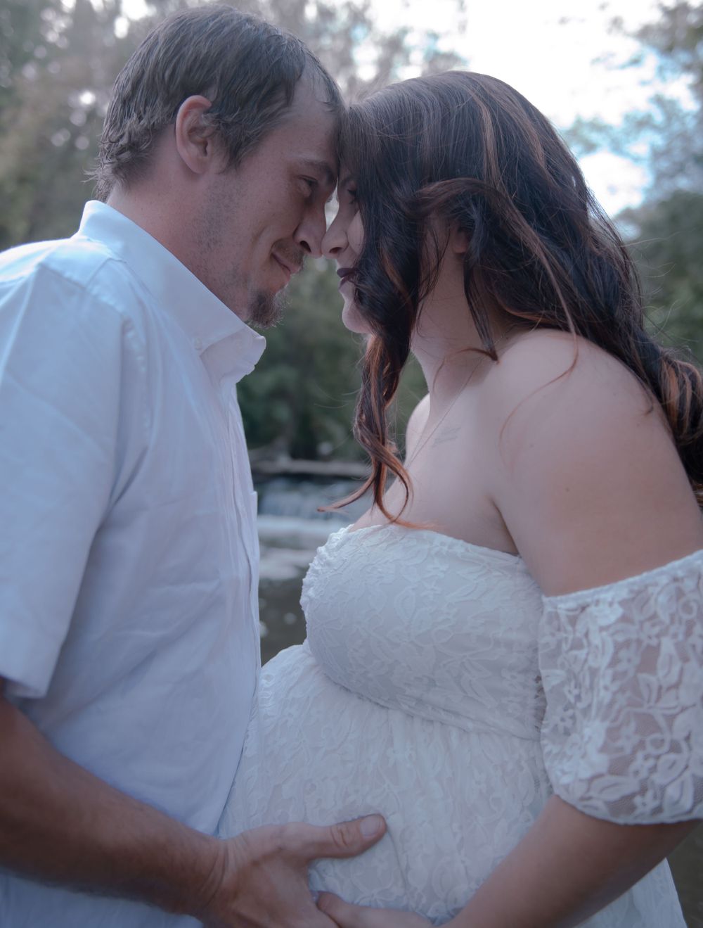 A tender maternity moment captured between a couple in white clothing by the water.