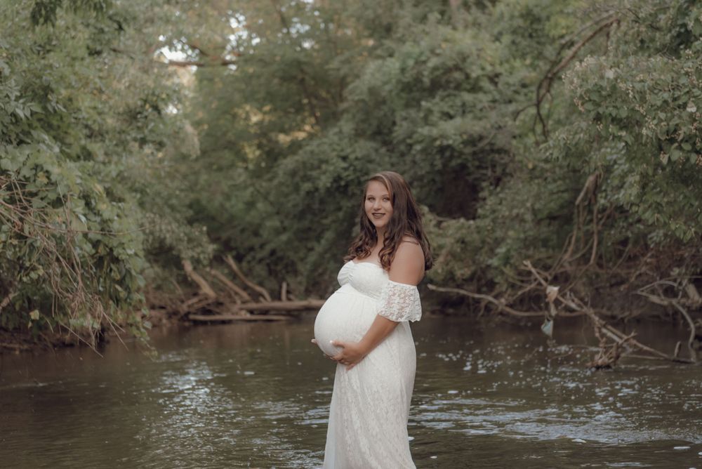 Maternity portrait by a stream wearing flowing white dress in natural setting.