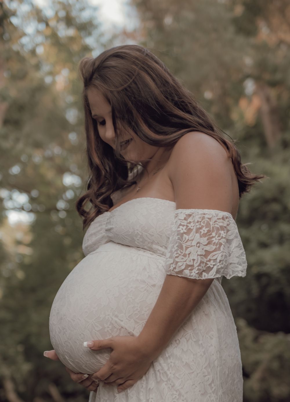 Expectant mother wearing off-shoulder white lace dress in outdoor nature setting.