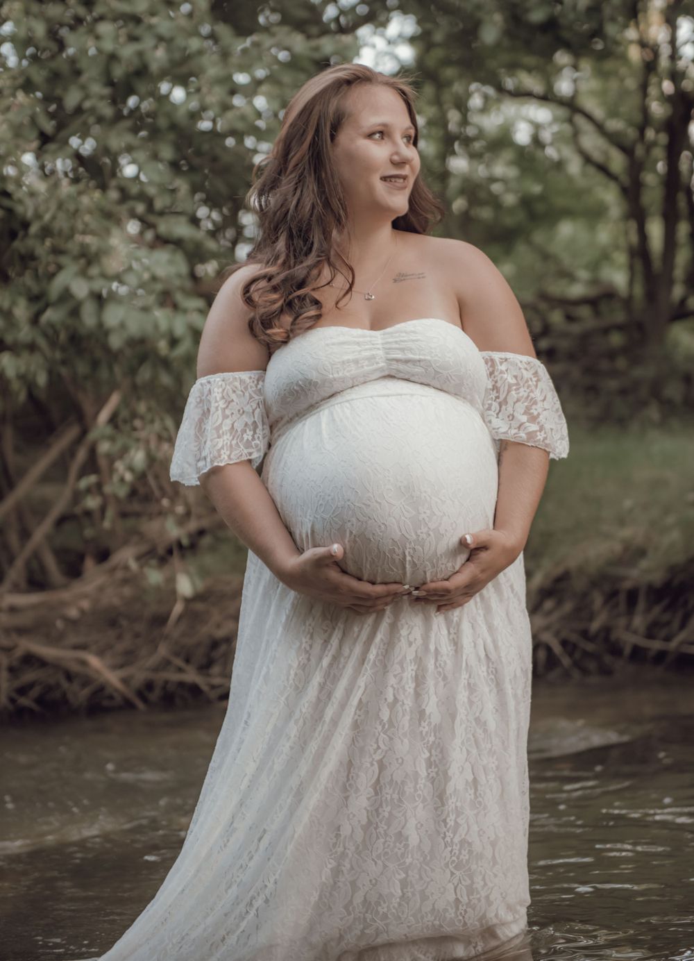 Elegant maternity portrait in white lace dress by water's edge.
